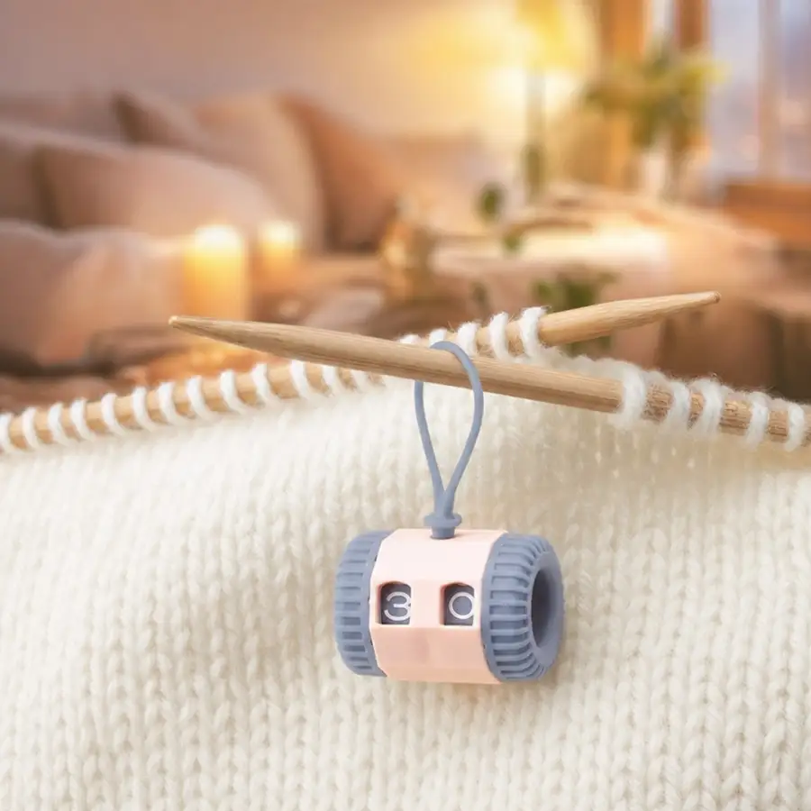 Pastel on-needle row counter riding a wooden needle above cream knitting, cozy living room behind